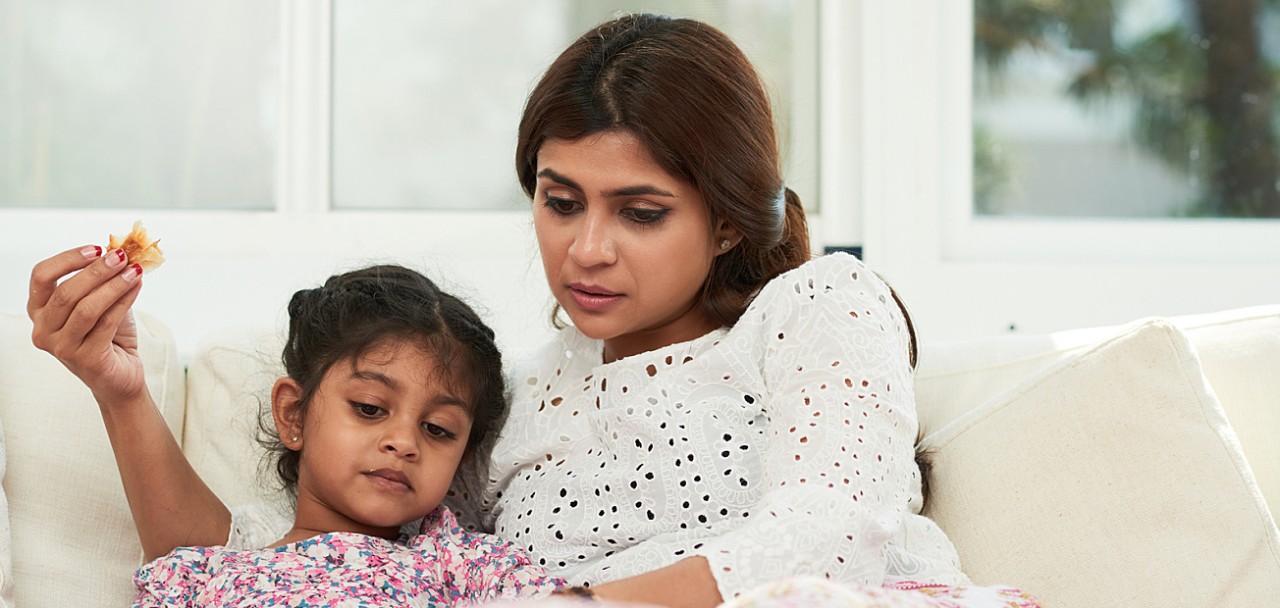 Spending lazy weekend at home: pretty Indian woman and her cute little daughter lying on cozy sofa, eating delicious pastry and watching cartoon on digital tablet