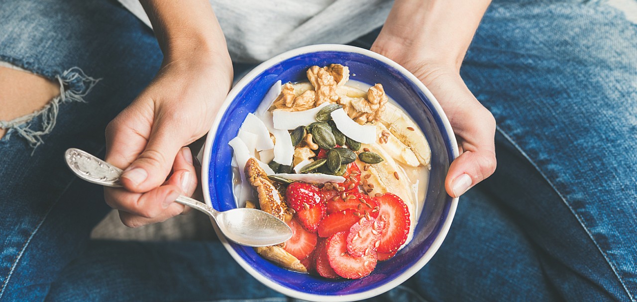 Eating healthy breakfast bowl. Yogurt, granola, seeds, fresh and dry fruits and honey in blue ceramic bowl in woman' s hands. Clean eating, dieting, detox, vegetarian food concept