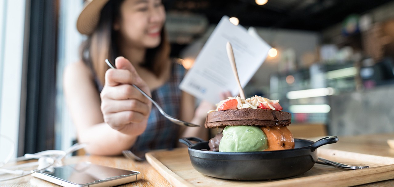 Happy asian woman's traveller wearing dress with brown hat sitting and eating ice cream with chocolate waffles ontop with almond and fruit serve on wooden plate in dessert café while reading a book.