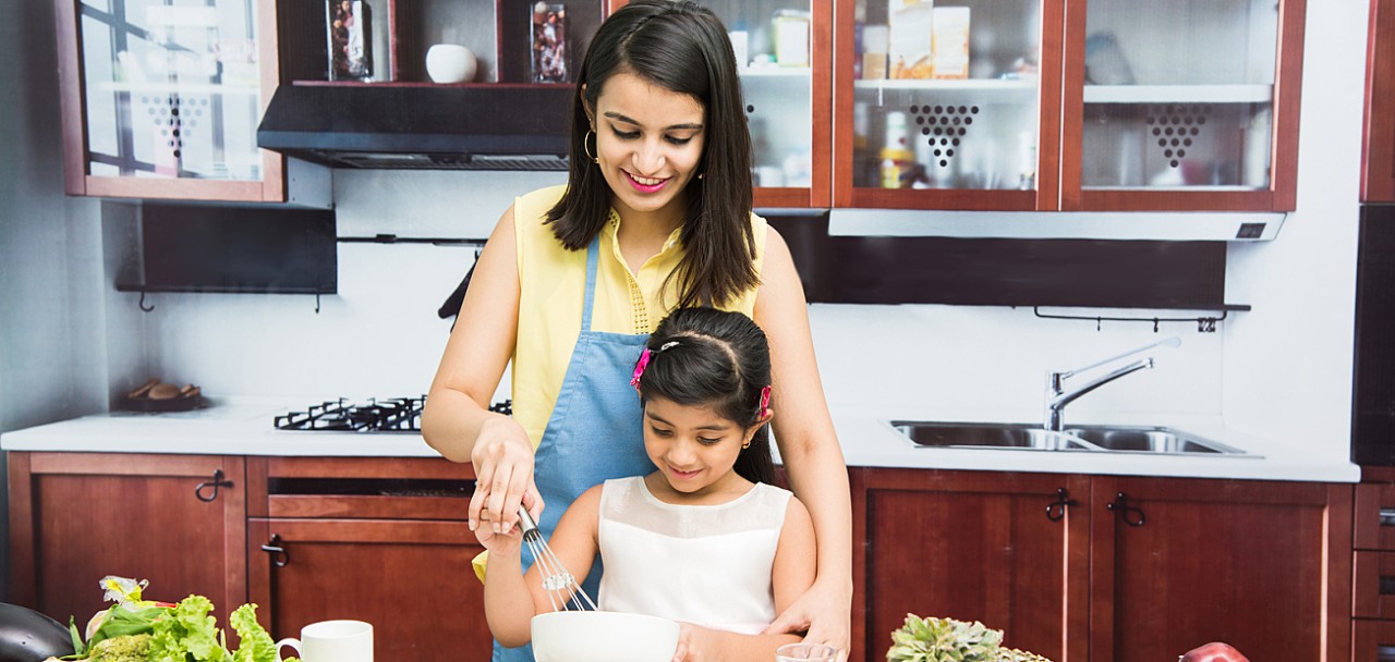 Pretty Indian young lady or mother with cute girl child or  daughter in kitchen having fun time with table full of fresh vegetables and fruits