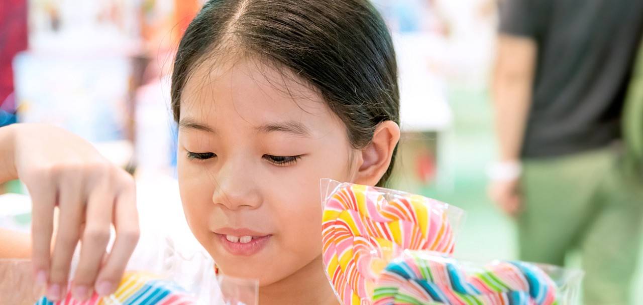 Happy Asian young girl buying colorful of sweets or lollipop in the store with blurred people background. Pretty child with candy unhealthy eating concept. Merry Christmas and happy new year day.