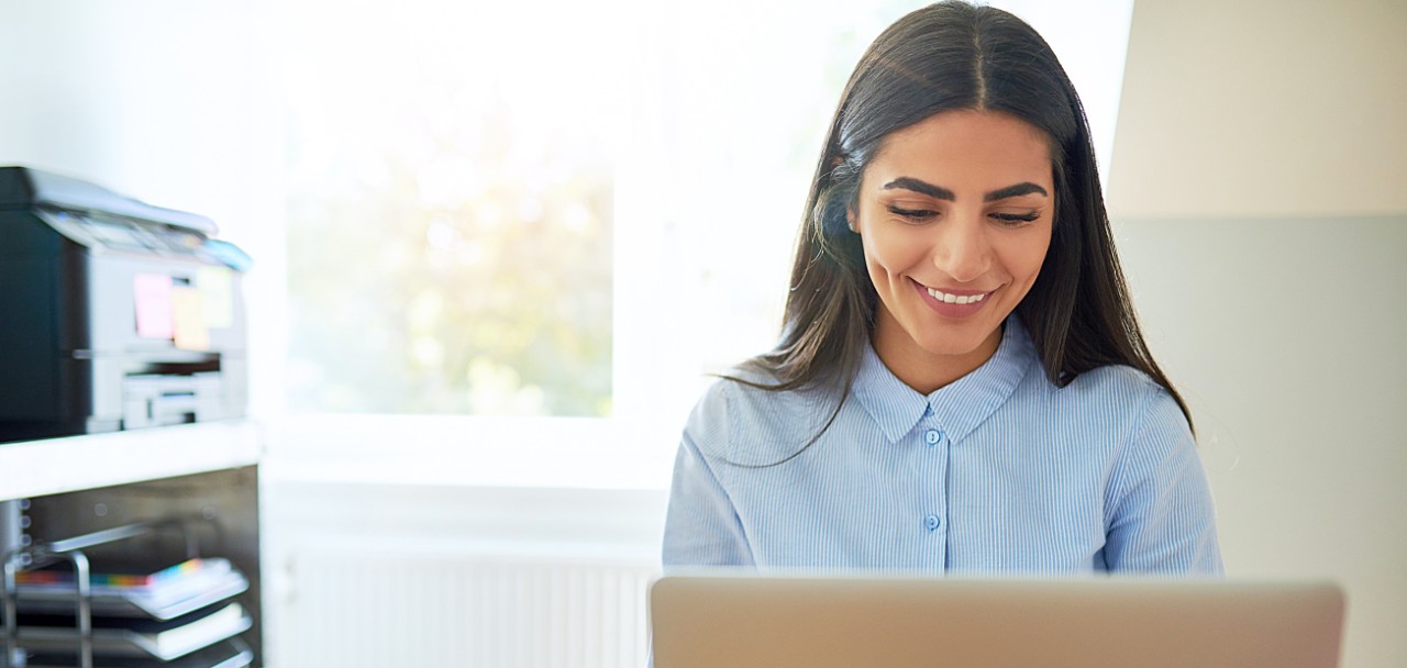 Single laughing young Indian woman beside printer on shelf and wearing long hair while seated in front of laptop computer in bright room; Shutterstock ID 507957673; PO: 123