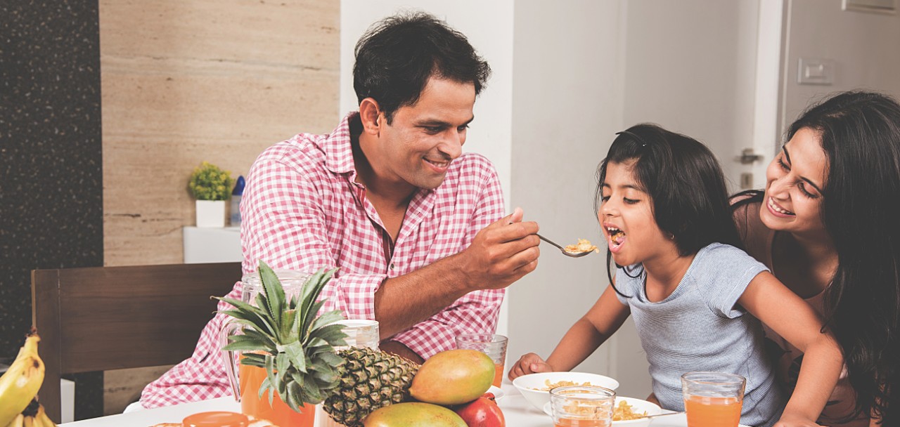 An attractive happy, smiling Asian Indian family of mother, father and daughter. Father feeding cereal to daughter with spoon at dining table. Indians eating breakfast, lunch / dinner. Selective focus; Shutterstock ID 667261060; PO: 123
