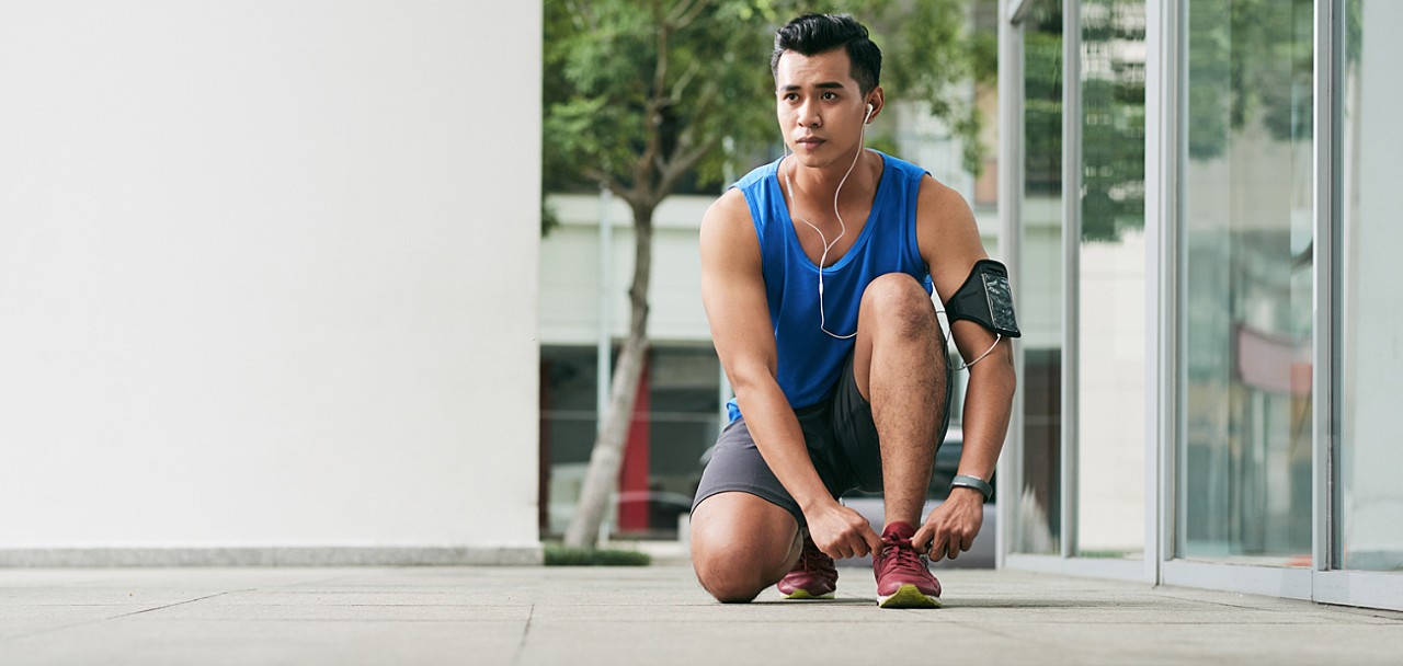 Portrait of young Asian sportsman listening to music in headphones while tying his shoelaces before morning run