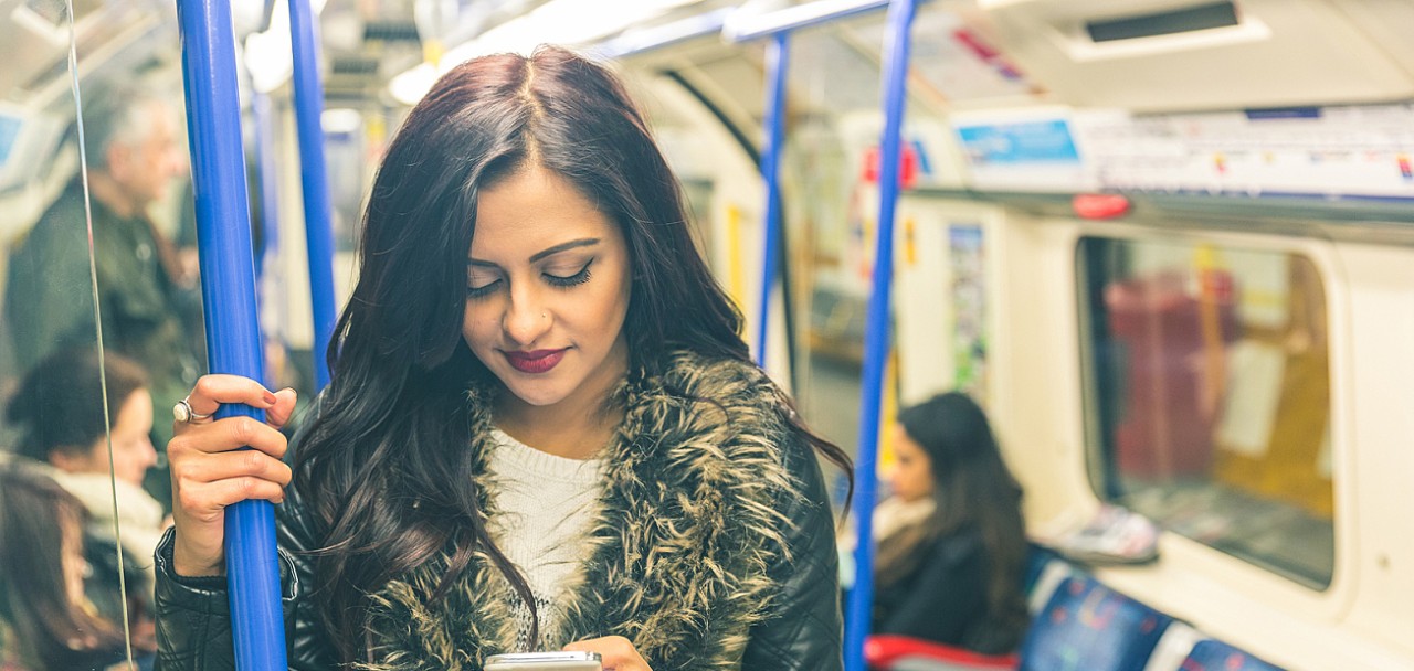 Young indian woman looking at her smart phone and typing while travelling on the tube in London. She is standing and holding with an hand. Transport and commuting concepts. Colours filter added.