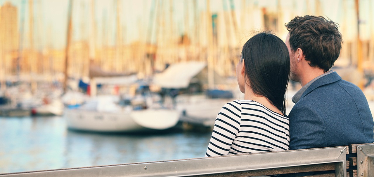 Lovers couple Romantic dating sitting on bench on date in old harbour, Port Vell, Barcelona, Catalonia, Spain. Happy woman and man embracing enjoying life and romance outside