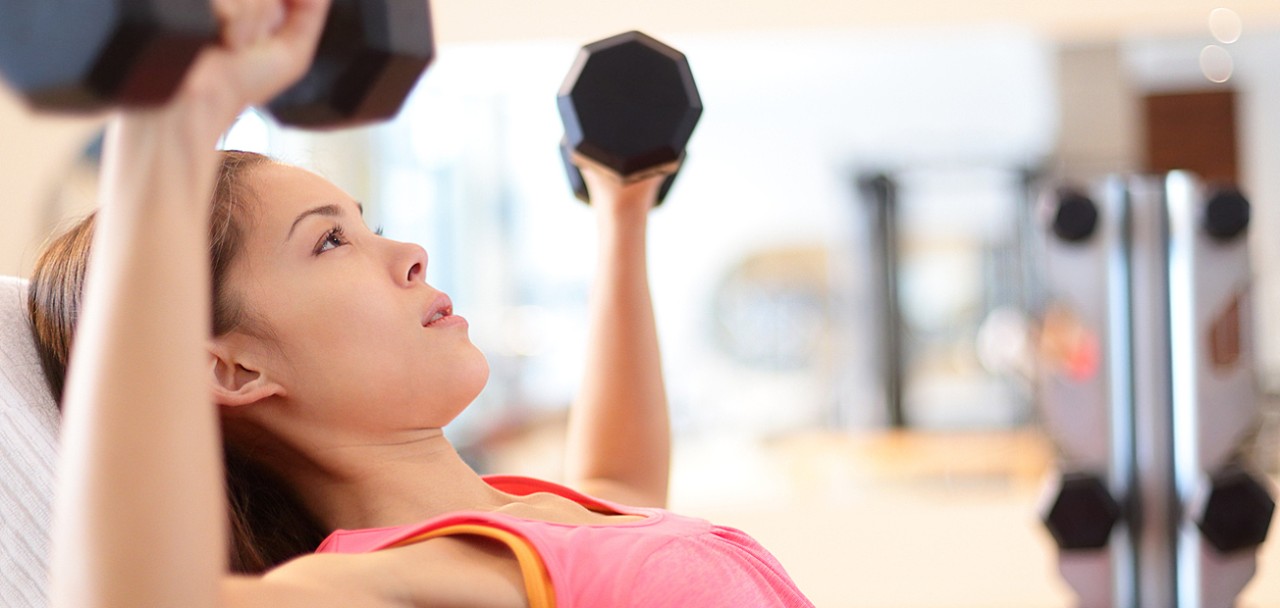 Gym woman strength training lifting dumbbell weights in shoulder press exercise. Female fitness girl exercising indoor in fitness center. Beautiful fit mixed race Asian Caucasian model training.