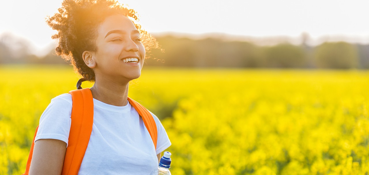 Outdoor portrait of beautiful happy laughing mixed race African American girl teenager female young woman with drinking water bottle in a field of yellow flowers at sunset in golden evening sunshine; Shutterstock ID 672089662; PO: 123